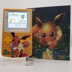 Puede incluir: Una consola Game Boy Color amarilla con Eevee de Pokémon. La pantalla muestra una escena de batalla con Pikachu. La consola y la ilustración de la caja muestran a Eevee. La consola tiene un D-pad y botones amarillos, con el logo de Pokémon.