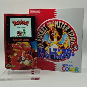 May include: A red transparent Game Boy Color handheld console featuring a Charizard graphic, displayed on a clear stand next to its original box. The screen shows the Pokémon Red Version title screen with a character and a Pidgey sprite.