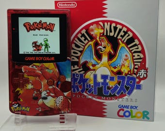 May include: A red transparent Game Boy Color handheld console featuring a Charizard graphic, displayed on a clear stand next to its original box. The screen shows the Pokémon Red Version title screen with a character and a Pidgey sprite.