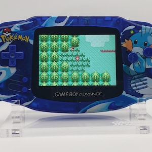 Puede incluir: Una consola portátil Game Boy Advance azul con temática Pokémon. La pantalla muestra una escena de juego pixelada. La consola tiene un diseño de olas azules y un gráfico del personaje Mudkip. El texto "GAME BOY ADVANCE" es visible.