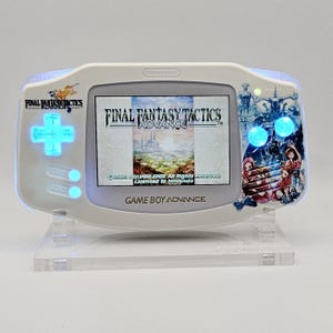 May include: A white Game Boy Advance handheld console featuring a Final Fantasy Tactics Advance theme. The device has a backlit screen displaying the game title, glowing blue buttons, and a character illustration on the right side of the casing.