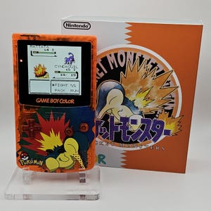 Può includere: Una console portatile Game Boy Color arancione trasparente con grafica di Cyndaquil e marchio Pokémon, esposta con la sua scatola arancione e bianca abbinata. Lo schermo mostra una battaglia tra Rattata e Cyndaquil in interfaccia pixel.