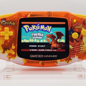 May include: A bright orange and yellow Game Boy Advance handheld video game console with a fiery design. The screen displays the Pokemon FireRed Version title screen. The console is on a clear plastic stand.