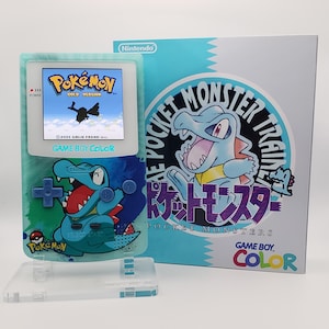 May include: A teal Game Boy Color console with a Pokémon Gold Version game displayed on the screen. The console features a design of a blue crocodile-like Pokémon. The box next to it has the text "POCKET MONSTER TRAIN" and "GAME BOY COLOR".