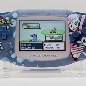 Op de afbeelding: Een blauwe, doorschijnende Game Boy Advance handheld console met een aangepast Pokémon-thema met Piplup en Dawn-afbeeldingen. Het scherm toont een gevecht tussen een Rattata en een Articuno, met de tekst What will ARTICUNO do? en menu-opties.
