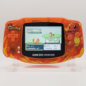 May include: Orange Game Boy Advance handheld console featuring a Pokemon theme. The screen displays a battle scene with Charmander. The console has a Pokemon logo and a Charmander graphic on the side.