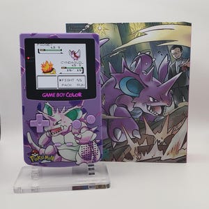 May include: A purple Game Boy Color handheld console featuring a Nidoking graphic, displayed on a clear acrylic stand. The screen shows a Pokémon battle between Pidgey and Cyndaquil. Behind the console is a matching box with Nidoking artwork.