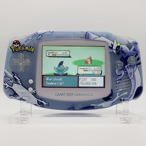 May include: A custom Game Boy Advance handheld console with a blue and white design featuring Pokémon characters. The screen displays a Pokémon battle scene. The console is on a clear stand.