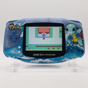 Puede incluir: Una consola portátil Game Boy Advance azul con temática Pokémon. La consola presenta una pantalla que muestra una escena del juego, una cruceta azul y un gráfico de Squirtle. La consola está decorada con gráficos de olas y Pokémon.