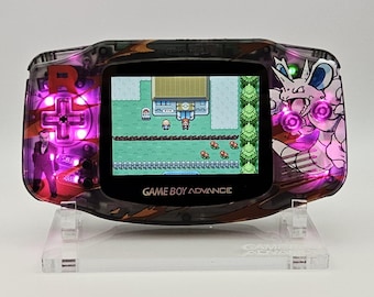 May include: A transparent black Game Boy Advance handheld console featuring a custom Team Rocket graphic with Giovanni and Nidoking. The device is illuminated by internal purple LEDs and displays a pixelated Pokémon game scene on the screen.