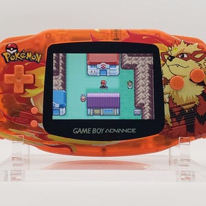 Può includere: Console portatile Game Boy Advance arancione con un design infuocato che presenta un personaggio Pokémon. Lo schermo mostra un gioco Pokémon con un personaggio in una città.