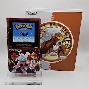May include: A red transparent Game Boy Color handheld console featuring a graphic of the Pokémon Entei on the front, displaying the Pokémon Gold Version title screen. It is positioned in front of a white and orange box with the Nintendo logo.