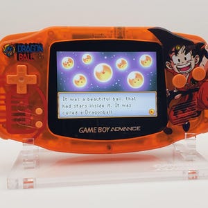 Puede incluir: Consola de videojuegos portátil Game Boy Advance naranja con un diseño de Dragon Ball Z. La pantalla muestra una escena del juego con el texto "It was a beautiful ball, that had stars inside it. It was called a Dragonball."