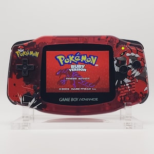 Puede incluir: Consola portátil Game Boy Advance roja con el juego Pokémon Versión Rubí en la pantalla. La consola tiene un diseño rojo y negro con gráficos de Pokémon. La pantalla muestra el título del juego y un gráfico de una criatura púrpura.