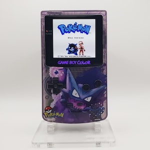 May include: A purple Game Boy Color handheld console featuring a Pokémon Blue Version game screen. The console has a purple and white design with a Pokémon character graphic. The screen displays the Pokémon logo and game details.