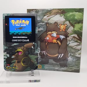 May include: A Game Boy Color console with a custom Pokemon design, displaying the "Pokemon Silver Version" game. The console is on a clear stand next to a matching box featuring a bear and fish illustration. The console has a clear shell.