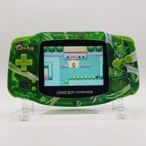 Puede incluir: Una consola portátil Game Boy Advance verde con temática Pokémon. La pantalla muestra una escena de juego con un edificio y personajes. La consola tiene una cruceta verde, botones y un logotipo de Pokémon. La consola está sobre un soporte transparente.