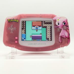 以下が含まれることがあります： 半透明のピンク色のゲームボーイアドバンス携帯ゲーム機。十字キーとボタンにはラメが入っています。画面にはピクセル化されたポケモンのゲームシーンが表示されています。本体右側にはピンク色のポケモン、ミュウのグラフィックがプリントされています。