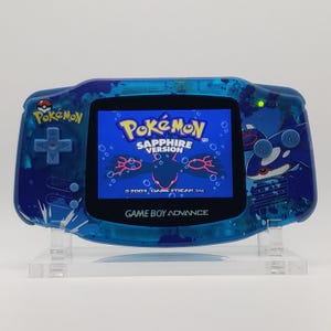 Puede incluir: Consola Game Boy Advance azul con el juego Pokémon Edición Zafiro. La consola portátil tiene una carcasa azul transparente con gráficos de Pokémon. La pantalla muestra el título del juego y el logotipo de Game Boy Advance.