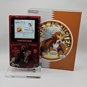 Op de afbeelding: Een rode, doorschijnende Game Boy Color handheld console met een afbeelding van Entei op de voorzijde, geplaatst op een doorzichtige standaard naast de originele doos. Het scherm toont een Pokémon-gevecht tussen Pidgey en Cyndaquil.