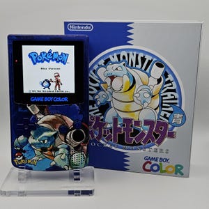 以下が含まれることがあります： カメックスのグラフィックが施された青い任天堂ゲームボーイカラー本体。画面には「ポケットモンスター 青」のタイトルが表示されている。隣には、同じくカメックスが描かれた対応するゲームソフトの箱が置かれている。