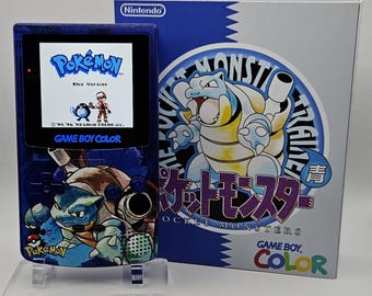 May include: A blue Nintendo Game Boy Color handheld console featuring a Blastoise graphic on the casing, displaying the Pokémon Blue Version title screen. Beside it stands the matching game box, which displays the Pokémon character Blastoise.