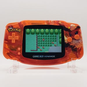 Puede incluir: Una consola portátil Game Boy Advance naranja con un tema de Pokémon. La pantalla muestra una escena de videojuego pixelada. La consola tiene una carcasa transparente y texturizada con el logotipo de Pokémon y un gráfico de personaje. El texto "GAME BOY ADVANCE" es visible.