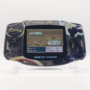May include: A Game Boy Advance handheld console with a custom design featuring a Pokémon theme. The screen displays a battle scene with Eevee and Diglett. The console has a transparent shell with black and gold accents.
