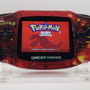 Puede incluir: Una consola portátil Game Boy Advance roja con un diseño personalizado que presenta un fondo rojo y negro ardiente con un cartucho de juego Pokemon Ruby Version insertado. La consola está en un soporte de acrílico transparente.