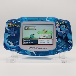 May include: A blue Game Boy Advance handheld console with a Pokemon theme. The console displays a battle scene with the text "Vaporeon" and "Pidgey". The console has a blue water-like design and the Pokemon logo.