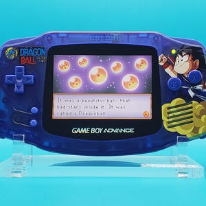 Könnte beinhalten: Transparente blaue Game Boy Advance-Konsole mit Dragon Ball-Grafiken. Der Bildschirm zeigt eine Szene mit leuchtenden Kugeln und Text. Die Konsole steht auf einem transparenten Ständer.