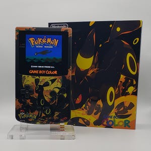 Peut inclure: Une console Game Boy Color noire avec un design Pokémon orange et noir personnalisé, avec le texte "Pokemon Silver Version" à l'écran. L'illustration de la boîte montre un personnage Umbreon noir avec des accents jaunes. La console est exposée sur un support transparent.