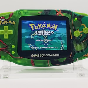 May include: A green Game Boy Advance handheld console with a Pokemon Emerald Version game cartridge inserted. The console has a green and black design with a red and orange pattern. The screen displays the game's title screen.