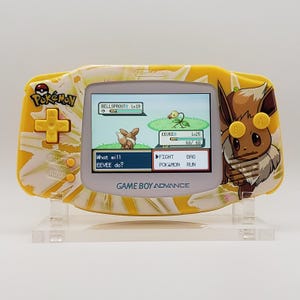 May include: A yellow Game Boy Advance handheld console with a Pokémon theme. The screen shows a battle scene with Eevee. The design incorporates a floral pattern and an image of Eevee. The text "Pokémon" and "Game Boy Advance" are visible.