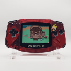 May include: A red Game Boy Advance handheld console displaying a video game scene. The console has a transparent red shell and features a directional pad, buttons, and a screen. The game on the screen shows a pixelated scene with a house and characters. The text "GAME BOY ADVANCE" is visible.