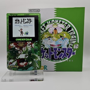 Puede incluir: Una consola portátil Game Boy Color verde translúcida con un gráfico de Venusaur en la parte frontal, mostrando la pantalla de título de Pocket Monsters Green Version. Está situada junto a una caja de juego verde y blanca a juego con Venusaur.