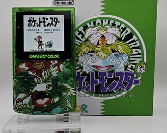 May include: A translucent green Game Boy Color handheld console featuring a Venusaur graphic on the front, displaying the Pocket Monsters Green Version title screen. It is positioned next to a matching green and white game box with Venusaur artwork.