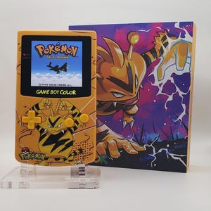 May include: A yellow Game Boy Color handheld console featuring an Electabuzz graphic and yellow buttons, displaying the Pokémon Gold Version title screen. Beside it stands a matching rectangular box with a large, colorful Electabuzz illustration.