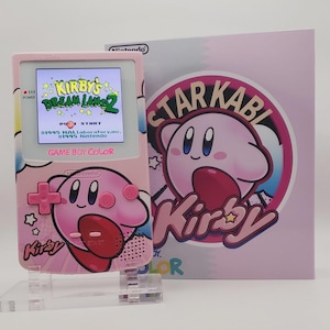 May include: A pink Game Boy Color handheld console featuring a Kirby graphic on the front, displaying the title screen for Kirby's Dream Land 2. Beside it stands a matching box with a large circular graphic of Kirby and the text Star Kabi Kirby.