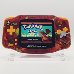 Puede incluir: Consola portátil Game Boy Advance roja y amarilla con el juego Pokémon FireRed Version. La consola tiene una carcasa roja transparente con gráficos de Pokémon y botones amarillos. La pantalla muestra la pantalla de título del juego.