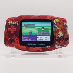 May include: A red Game Boy Advance console featuring a Pokemon theme. The screen shows a battle scene with the text "What will Totodile do?" and "GAME BOY ADVANCE". The console has a red and orange design with Pokemon characters.