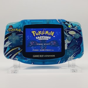 May include: A blue Game Boy Advance handheld video game console with a custom design featuring a blue and white wave pattern and a blue whale. The screen displays the Pokemon Sapphire Version game title.
