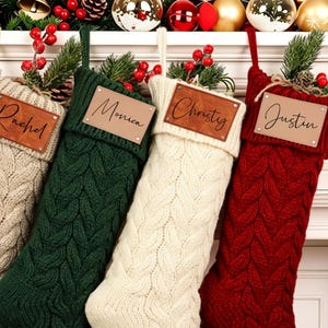 Può includere: Quattro calze natalizie in maglia a trecce nei colori beige, verde scuro, crema e rosso. Ogni calza presenta una targhetta rettangolare in pelle con i nomi Rachel, Monica, Christy e Justin incisi in nero, decorate con bacche e foglie verdi.