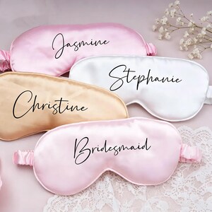 May include: Four satin sleep masks in light pink, white, and champagne colors. Each mask features a black script font name or title printed on the front: Jasmine, Stephanie, Christine, and Bridesmaid. The masks have elasticated side straps.