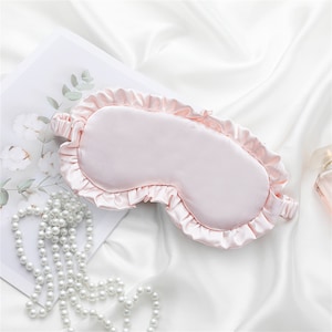 May include: A pink satin sleep mask with a ruffled edge. The mask is lying on a white surface.