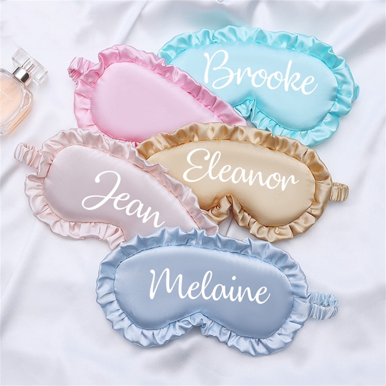 May include: Four satin sleep masks with ruffled edges in pink, beige, light blue, and blue. Each mask has a different name embroidered on it: Brooke, Eleanor, Jean, and Melanie.