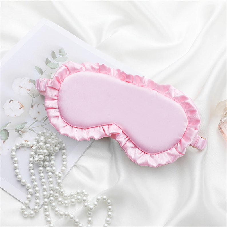 May include: A pink satin sleep mask with a ruffled edge. The mask is lying on a white surface.