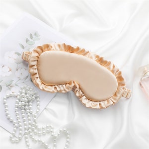 May include: A light beige silk sleep mask with a ruffled edge. The mask is lying on a white surface.