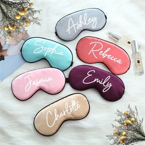 May include: Six personalized sleep masks in various colors, including pink, purple, blue, gray, and peach. Each mask has a different name written in white cursive lettering: Jasmin, Emily, Sophia, Ashley, Rebecca, and Charlotte.