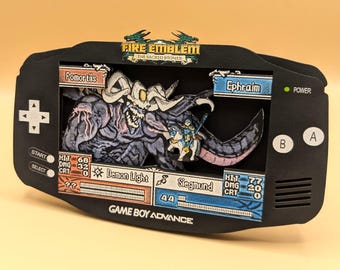 May include: A black Game Boy Advance shadow box display featuring a Fire Emblem: The Sacred Stones battle scene. The layered cutout shows Fomortiis and Ephraim with game stats, including Demon Light and Siegmund weapon text, in a 3D diorama style.